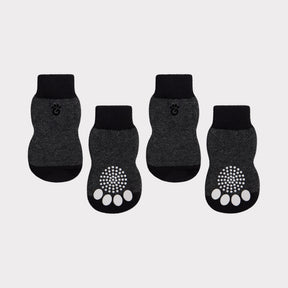 Four dark gray and black pet socks arranged on a light gray background, with two socks showing a white paw-print grip pattern on the sole and a small paw-print logo on the cuff.