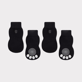 Four black pet socks arranged in a row on a light gray background. Two socks show the top view with a small paw print logo near the cuff, and two show the bottom view with white paw pad grip pads on the sole.